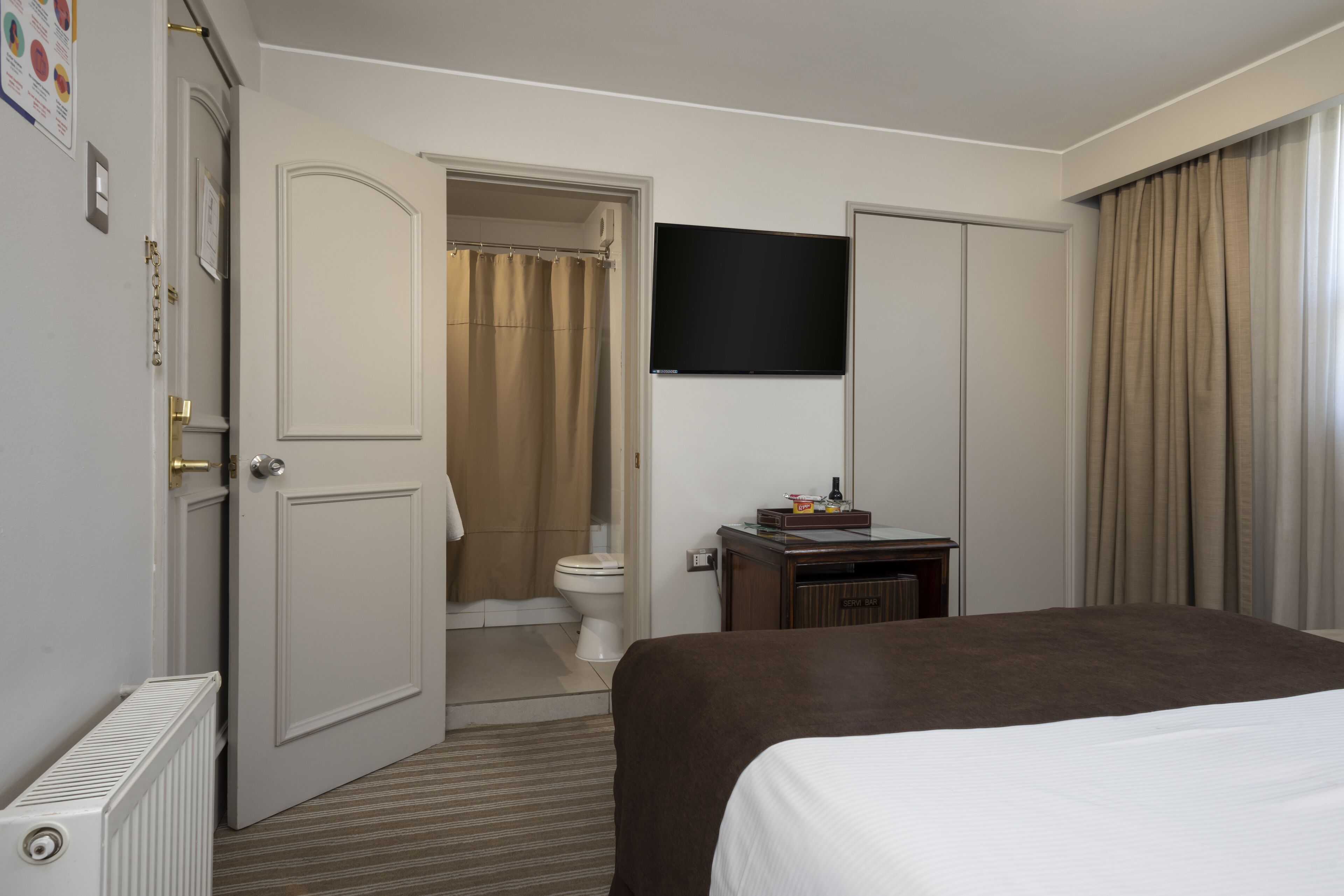 Une chambre d’hôtel équipée d’un lit, d’une télévision, d’une salle de bain avec des toilettes et un rideau de douche, ainsi que d’un radiateur.