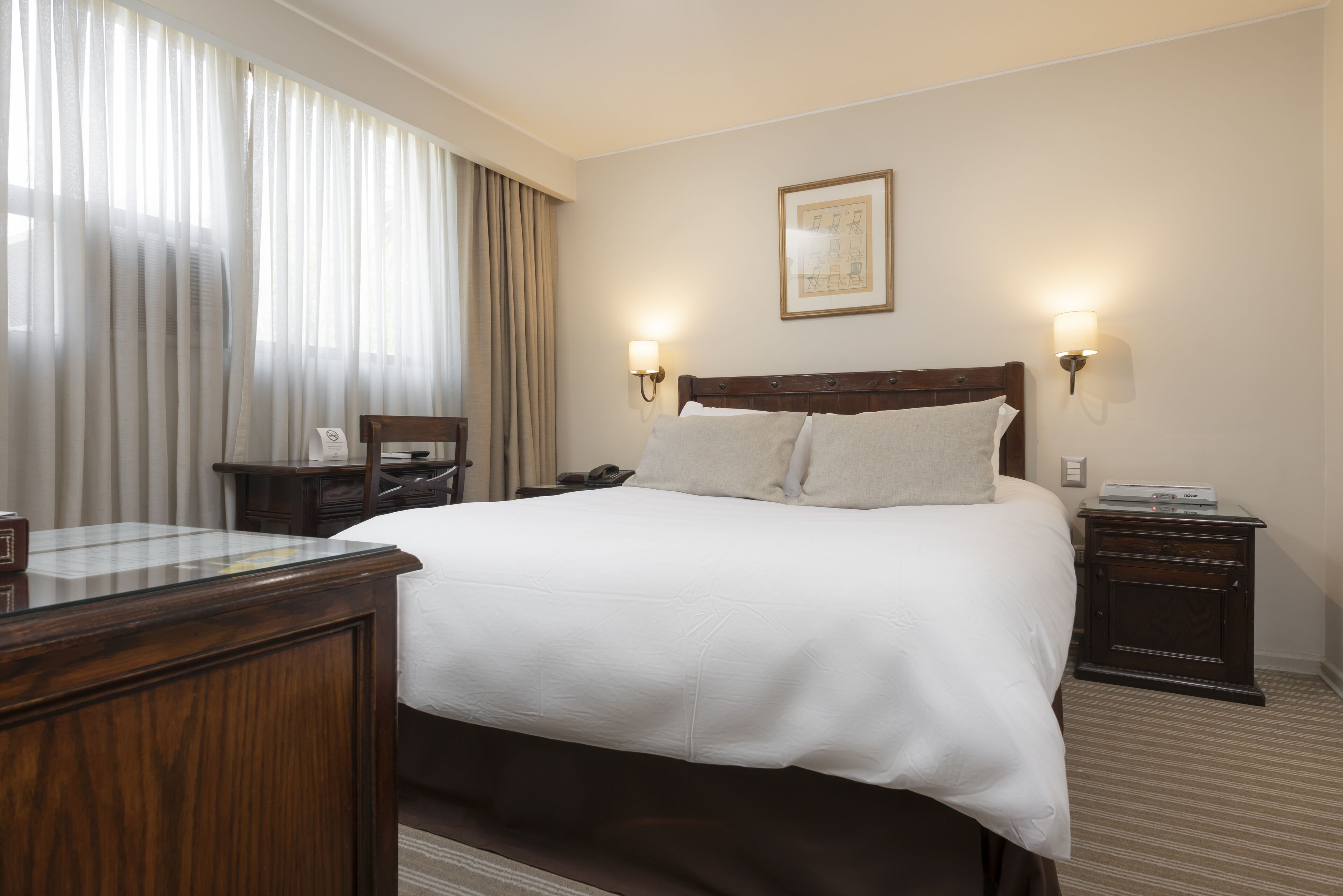 Ein Hotelzimmer mit einem großen Bett, zwei Nachttischen, einem Holztisch, einem Stuhl und einem Fenster mit Vorhängen.