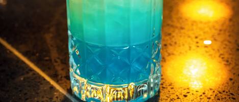 A tall glass with a gradient color, ice cubes, and a slice of orange on a bar counter.