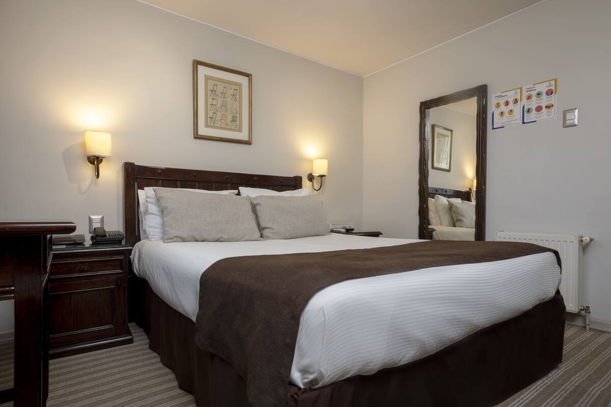 Ein Hotelzimmer mit einem großen Bett, Nachttischlampen, einem Holznachttisch, einem Telefon und einem Spiegel.