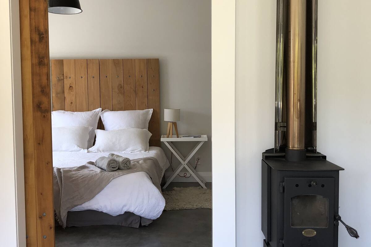 A bedroom with a wooden headboard, a bed with white linens, a bedside table with a lamp, and a wood-burning stove.