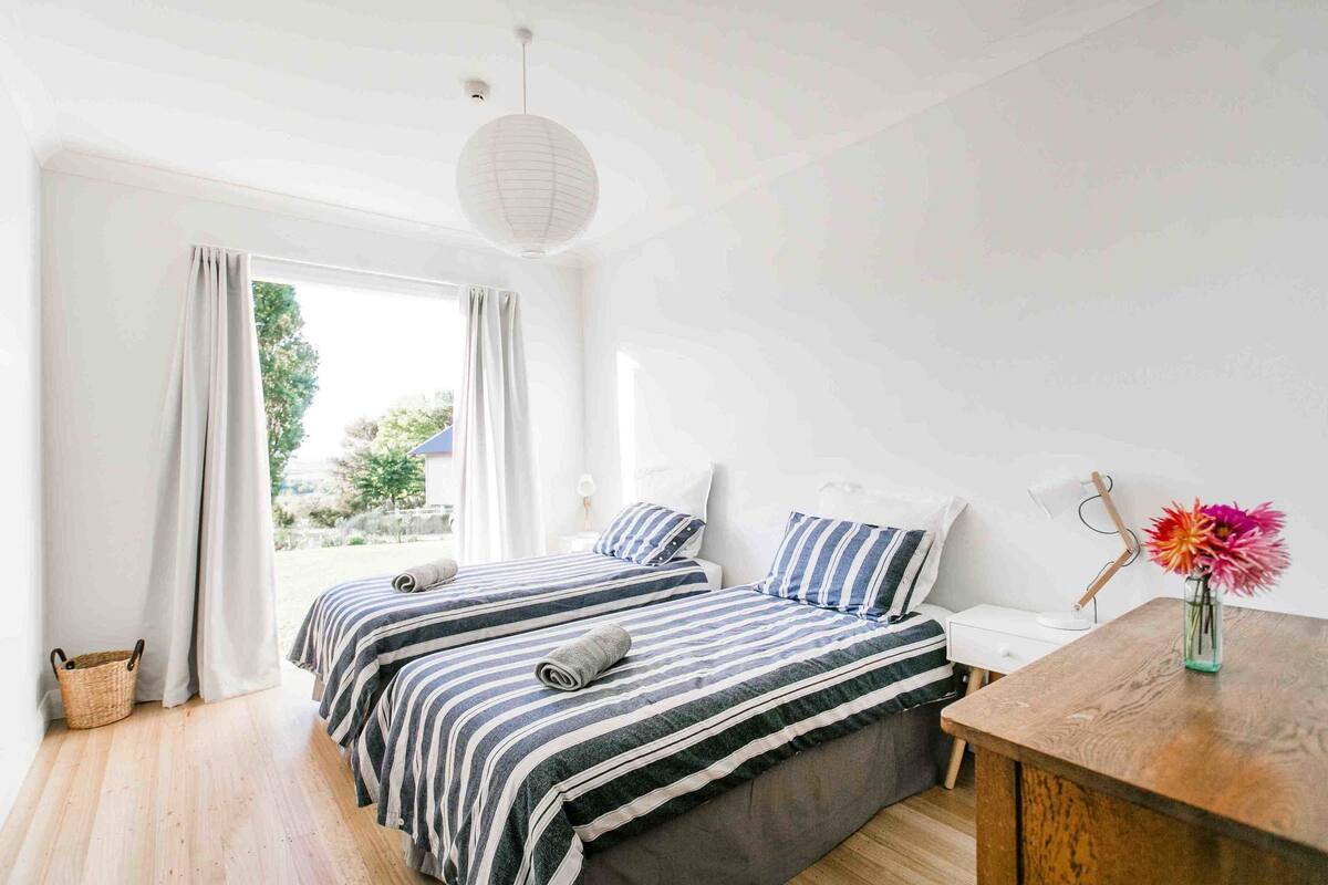A bedroom with two beds, a wooden desk, a lamp, and a vase with flowers.