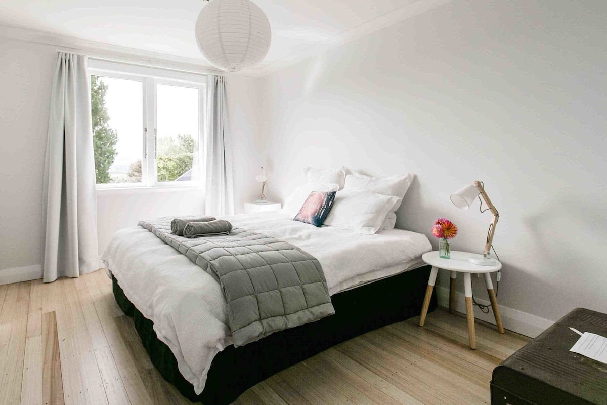 A bedroom with a bed, a bedside table with a lamp, a small table with a vase, and a window with curtains.