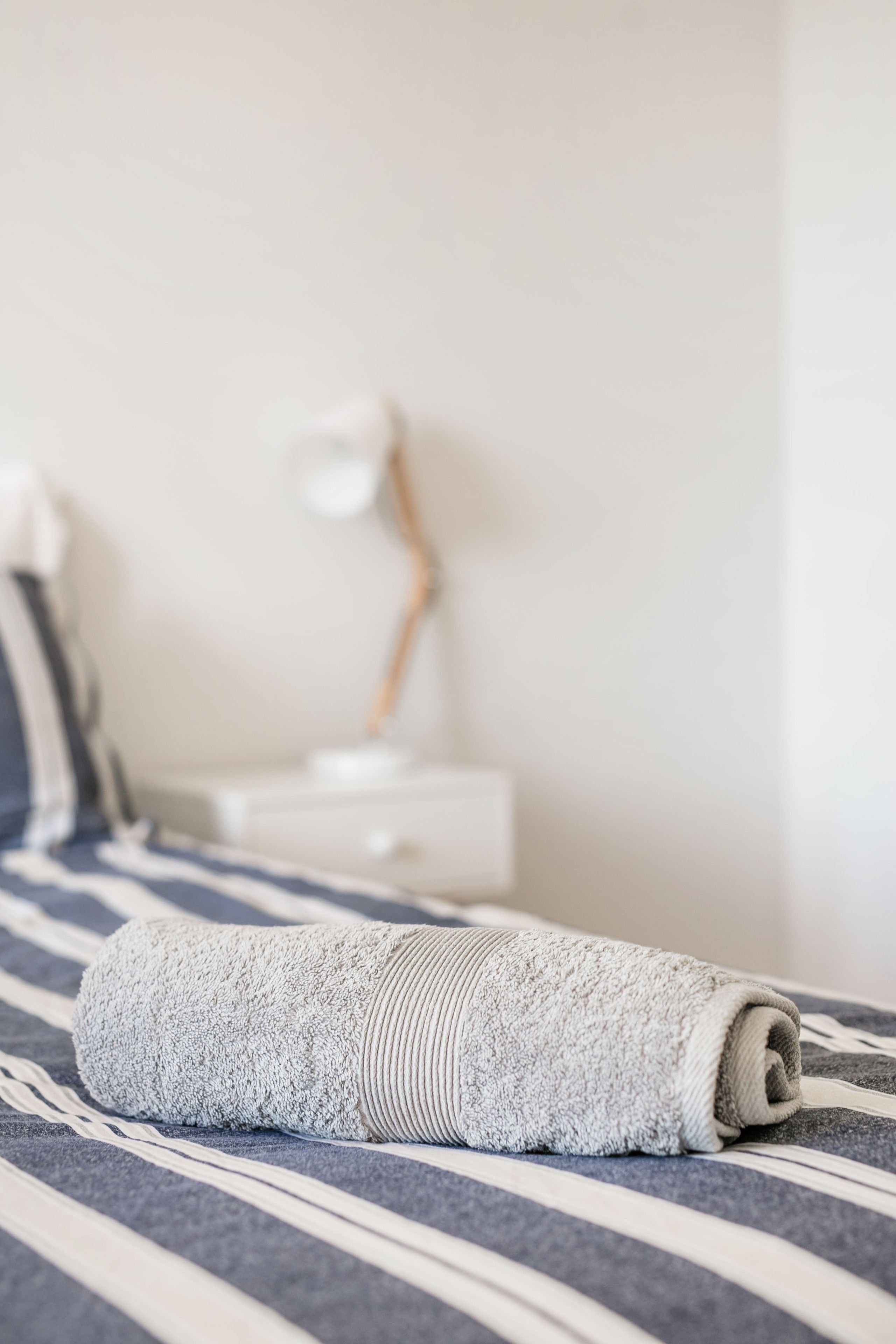 A neatly made bed with a striped bedspread, a rolled-up towel, and a bedside table with a lamp.