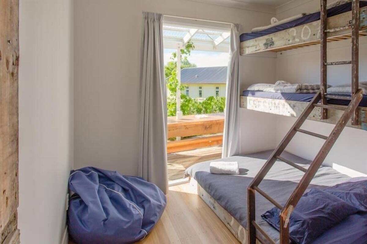 A dormitory room with bunk beds, a wooden ladder, a window with curtains, and a view of greenery outside.