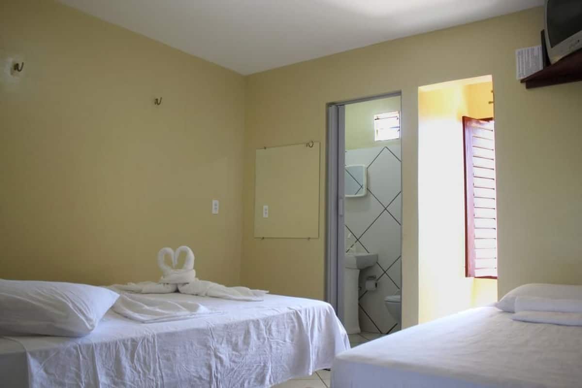 Una habitación de hotel con dos camas, un baño con puerta de vidrio y una ventana con persianas.