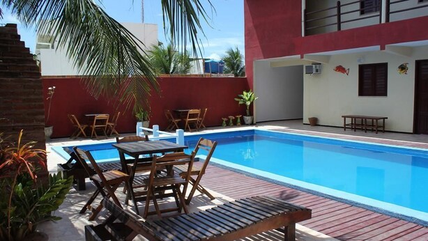 Una piscina con sillas de descanso y una mesa, rodeada de macetas con plantas y un muro rojo.