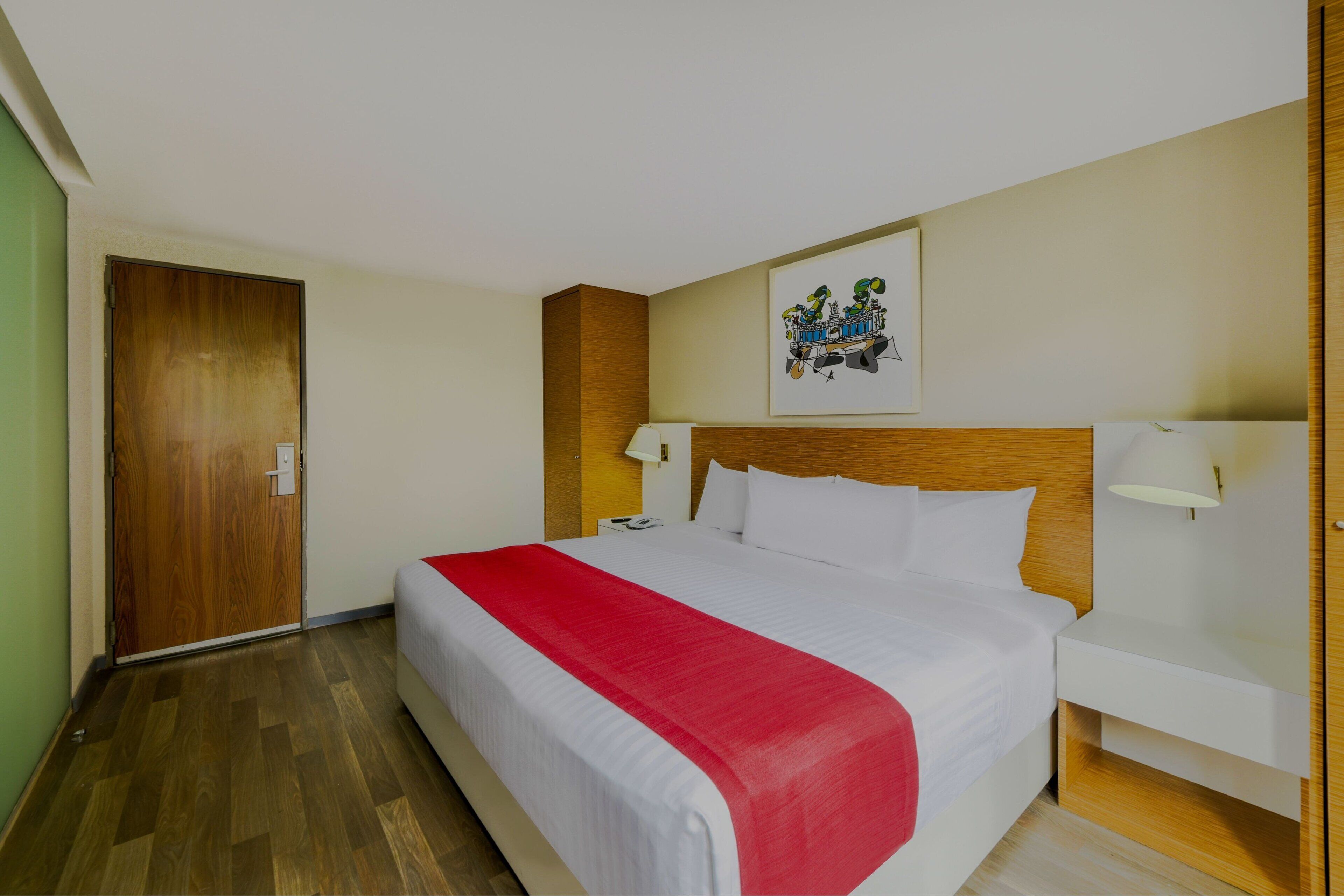 Hotelkamer met een houten hoofdbord, een groot bed met wit bedlinnen en een rode accentkleur, een nachtkastje met een lamp, en een deur links.