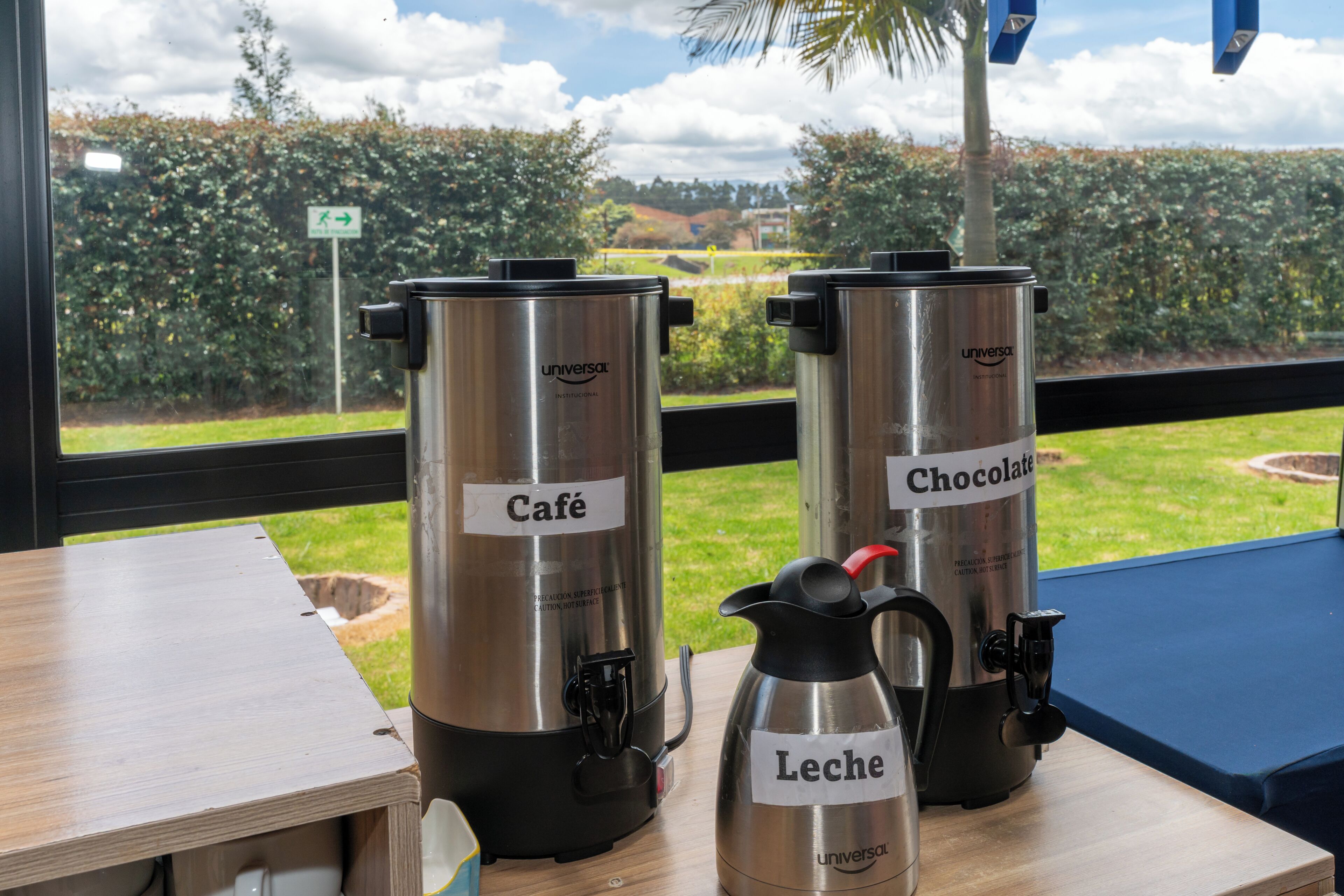 Two stainless steel coffee makers labeled 'Café' and 'Chocolate' on a table with a view of a garden through a window.