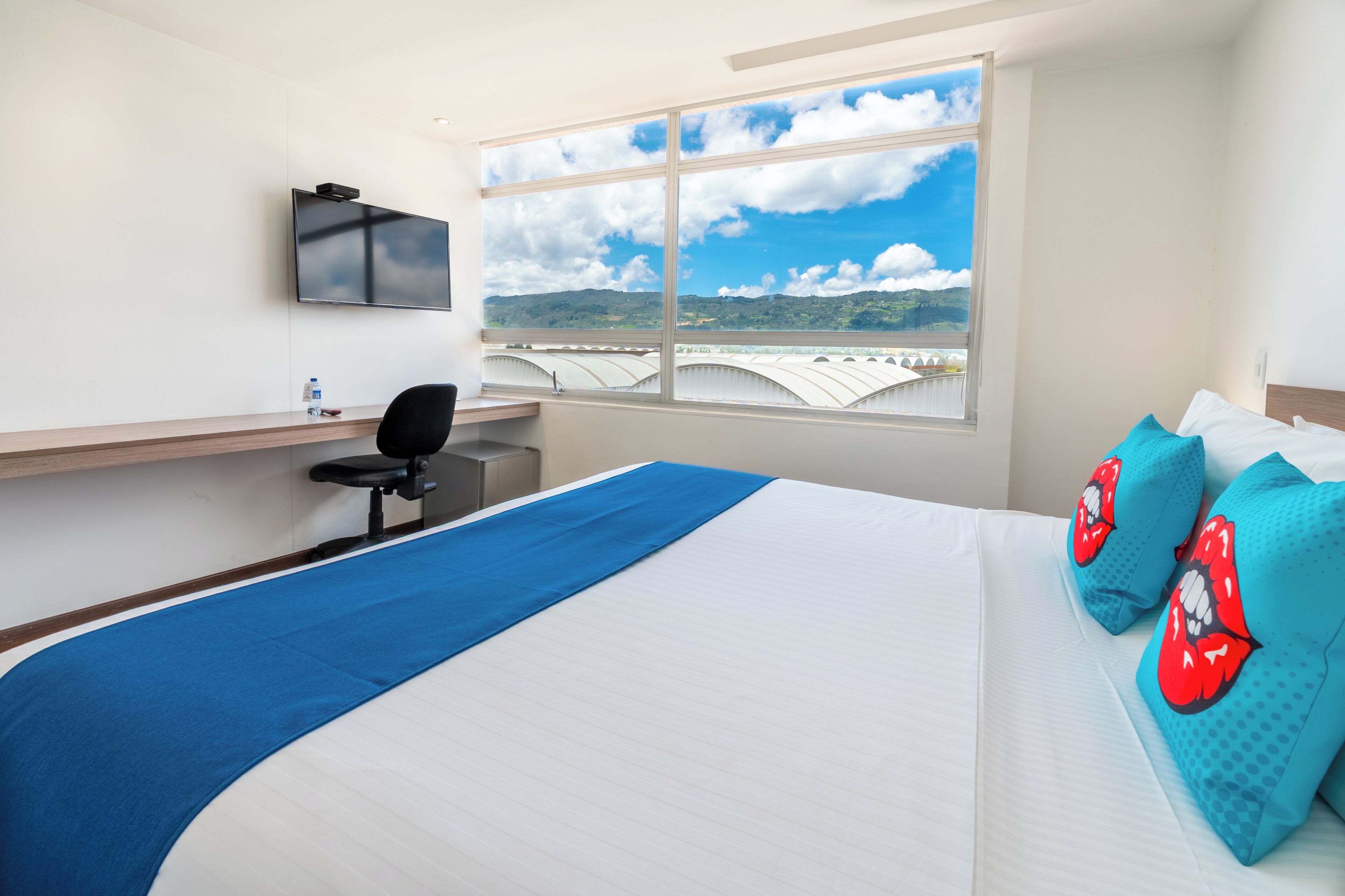 Une chambre d’hôtel équipée d’un lit, d’un bureau, d’une chaise, d’une télévision et d’une fenêtre donnant sur un paysage.
