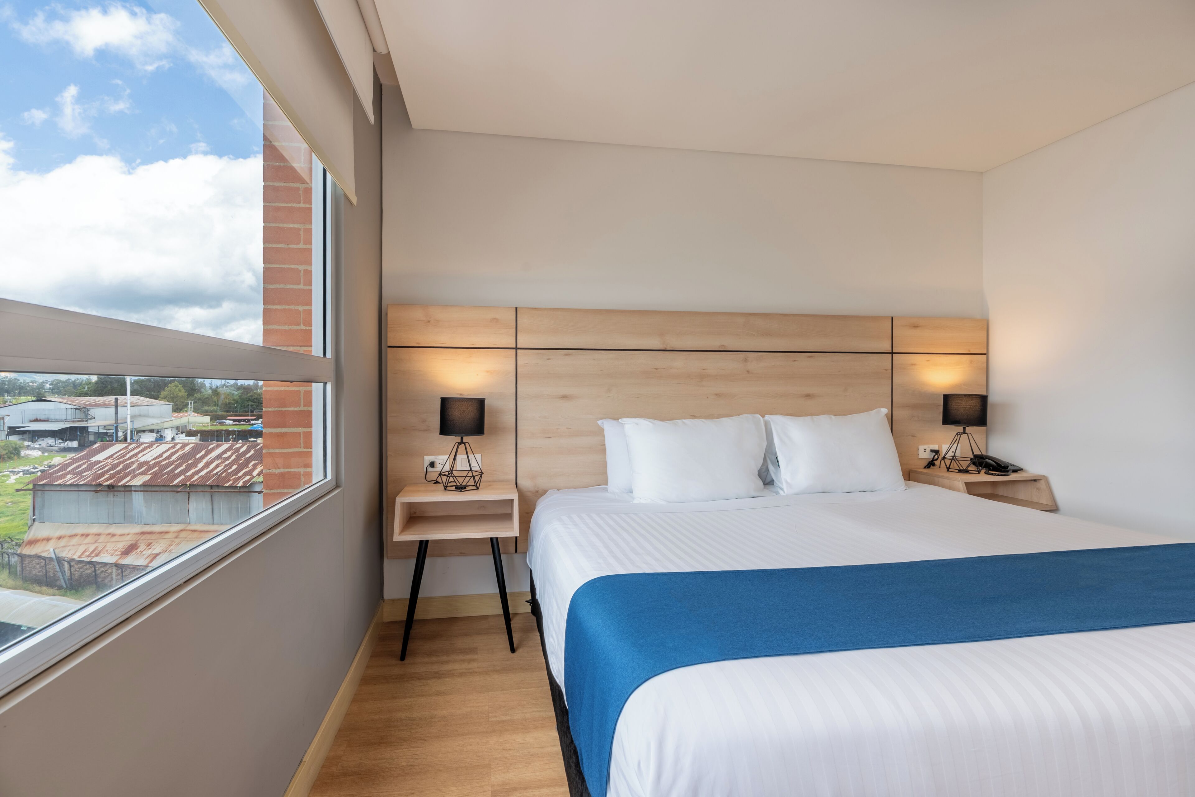 A hotel room with a large bed, a wooden headboard, two bedside tables with lamps, and a view of the outside through a large window.