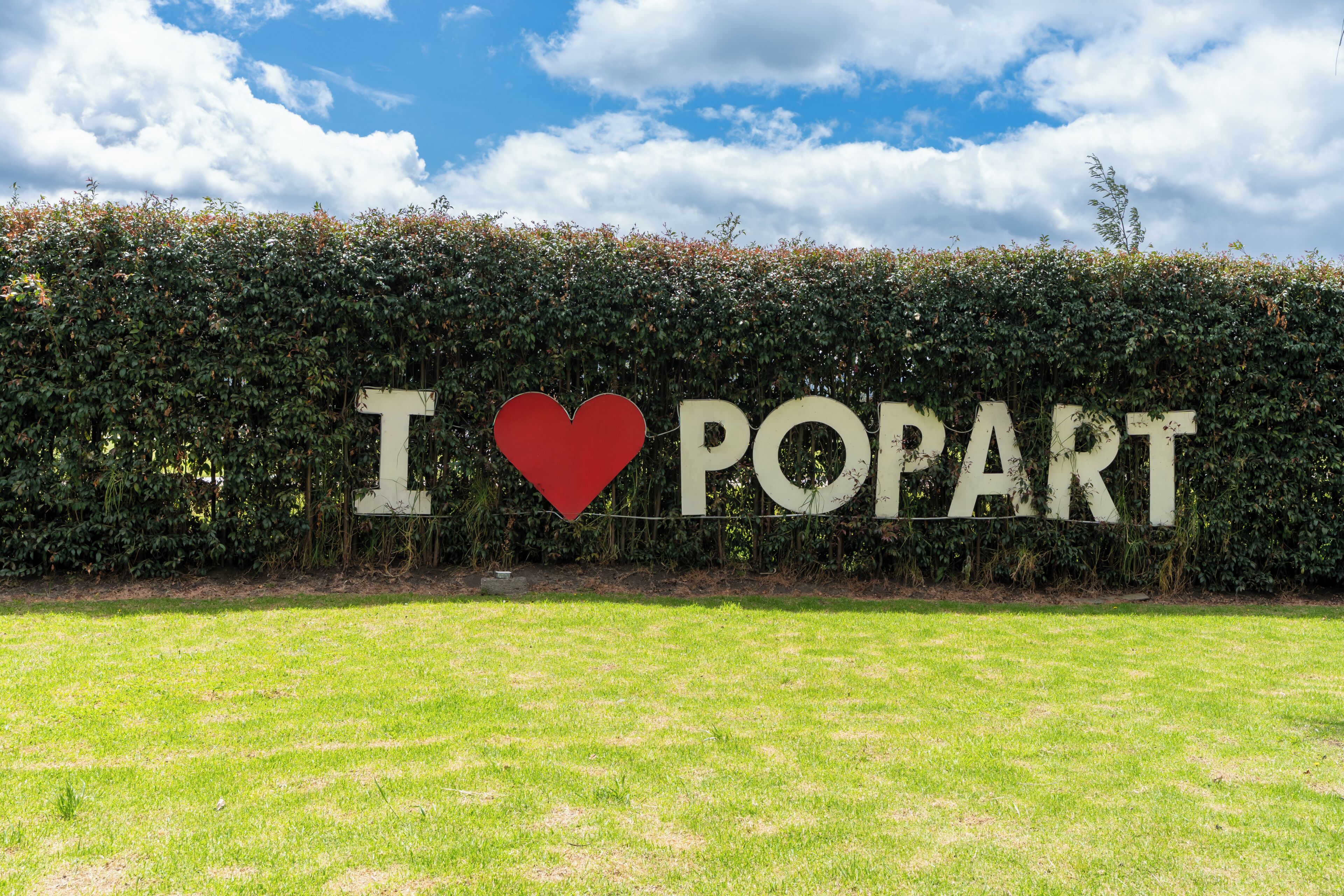 A sign with the text "I ♥ POPART" against a backdrop of a hedge and a clear sky.