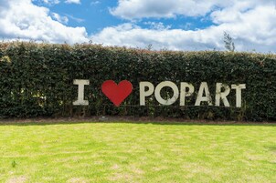 Un letrero con el texto "I ♥ POPART" sobre un fondo de seto y cielo despejado.