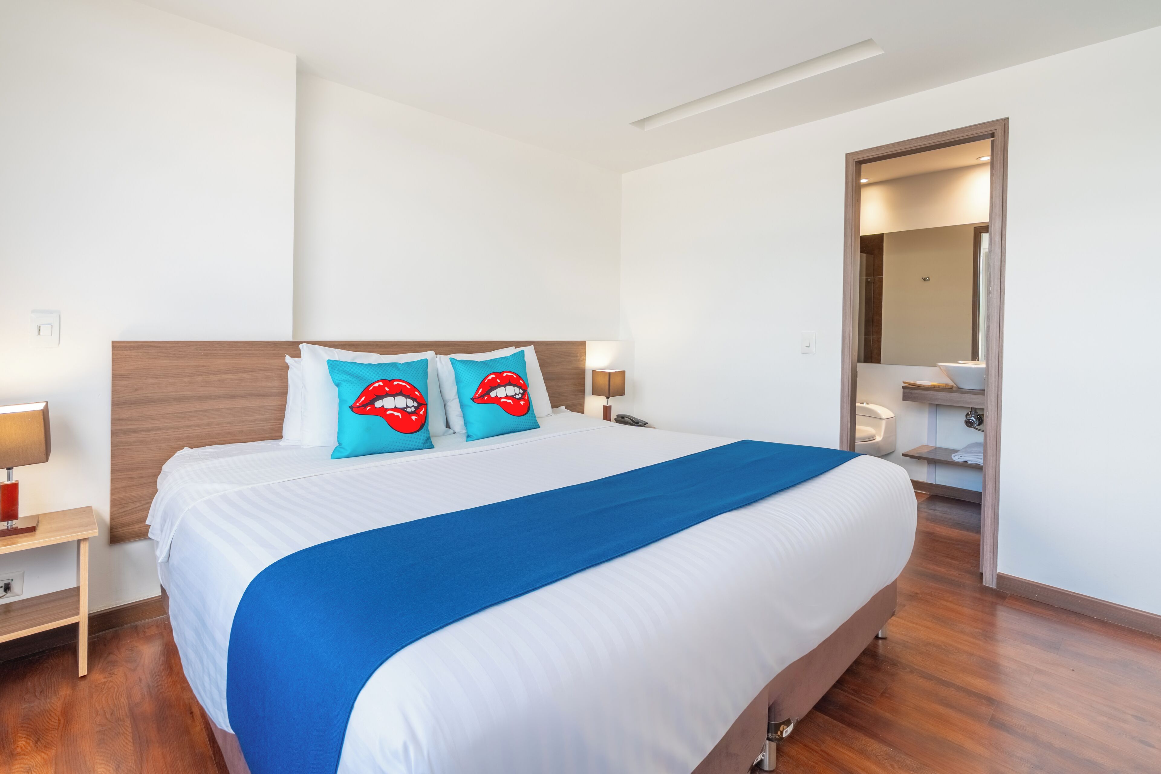 A hotel room with a large bed, wooden headboard, two bedside lamps, a blue bedspread, and a bathroom visible through an open door.