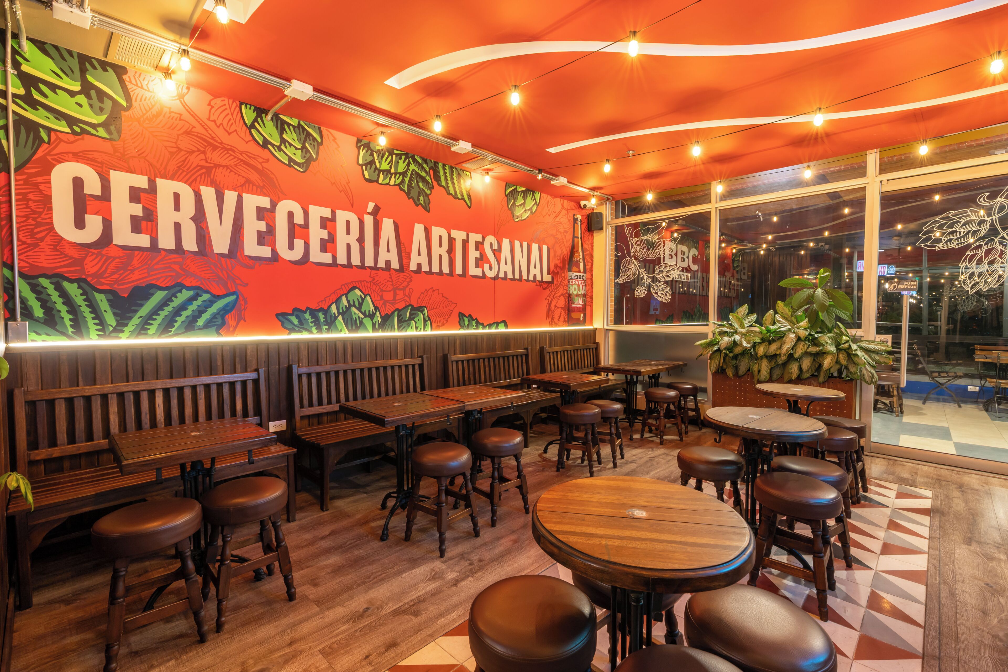 A restaurant interior with wooden tables and benches, a mural with the text "CERVEERÍA ARTESANAL," and large windows allowing natural light.