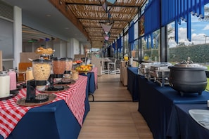 A buffet setup with various food items and beverages, a checkered tablecloth, and a view of the outdoors through large windows.