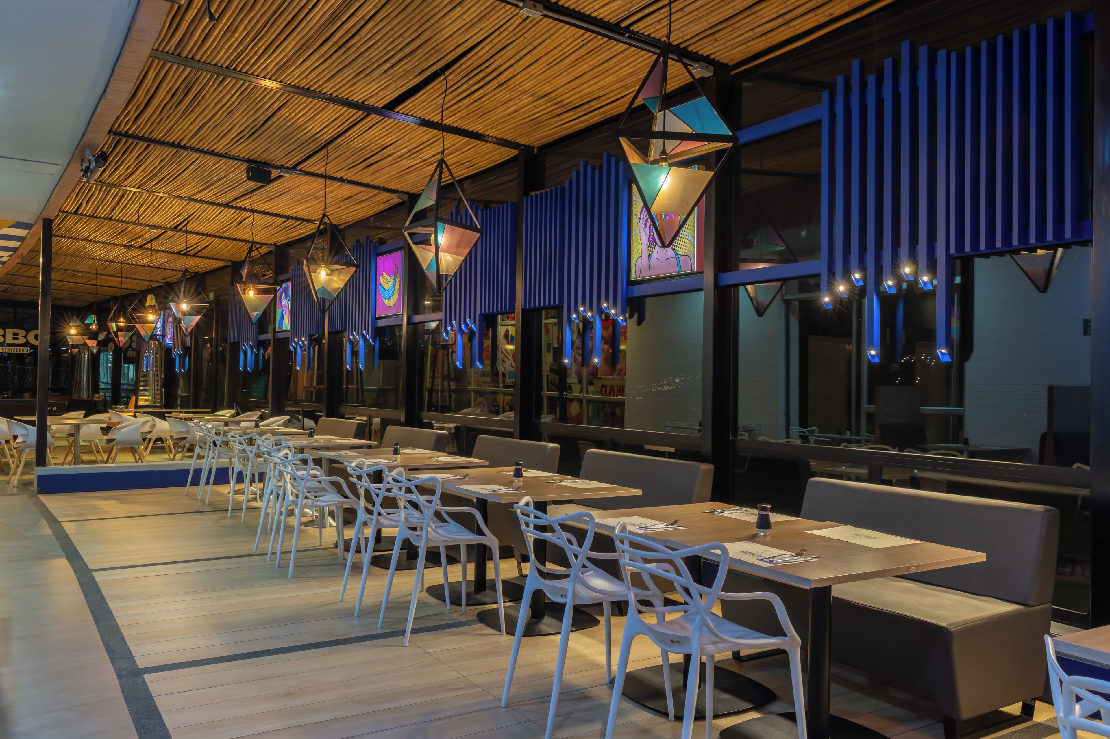 A restaurant interior with long tables and white chairs, a high ceiling with a bamboo-like design, and colorful hanging lights.