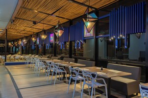 Interior de restaurante con mesas largas y sillas blancas, techo alto con diseño similar al bambú, y luces colgantes de colores.