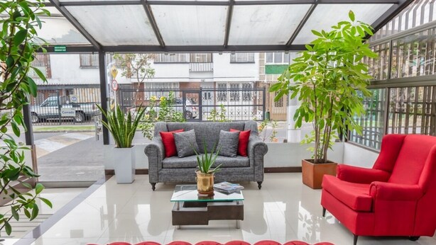 A modern indoor space with a grey sofa, red armchair, and a red tufted ottoman. There are potted plants and a glass coffee table. - Casa Quinta Hotel Embajada (Bogotá)