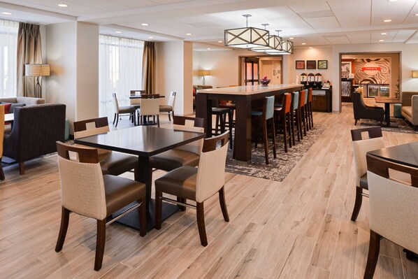 A hotel dining area with a long communal table, several chairs, a bar area, and a seating area with couches and a lamp.