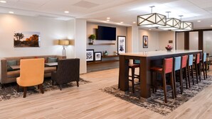 A modern hotel lobby with a wooden floor, a sofa, chairs, a bar area with stools, and a flat-screen TV mounted on the wall. - Hampton Inn El Centro (El Centro)