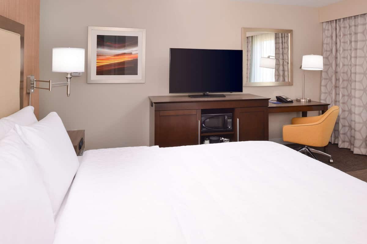A hotel room with a large bed, a desk with a flat-screen TV, a chair, and a desk lamp.