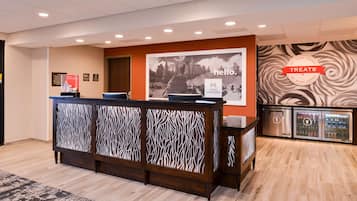 A hotel reception area with a wooden reception desk, a 'hello' sign, a 'Treats' sign, and a refrigerator with beverages.