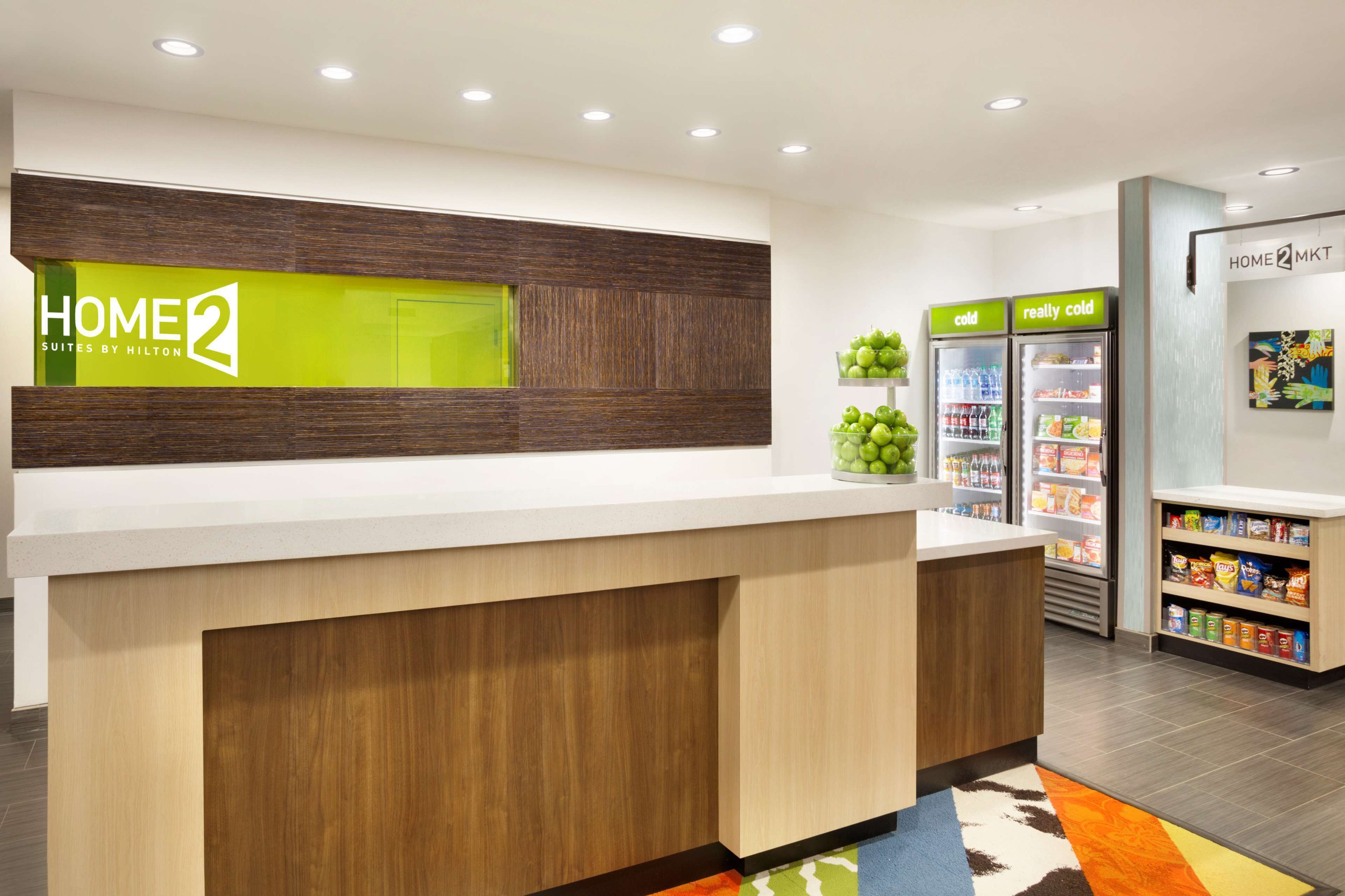 Un'area reception di un hotel dal design moderno, con una scrivania reception, un espositore di frutta e un refrigeratore per bevande.
