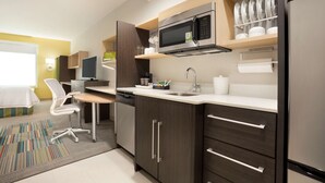 A modern kitchen with dark cabinetry, a stainless steel refrigerator, a microwave, and a dishwasher. There is a small dining area with a chair and table. - Home2 Suites by Hilton Houston Webster (Webster)