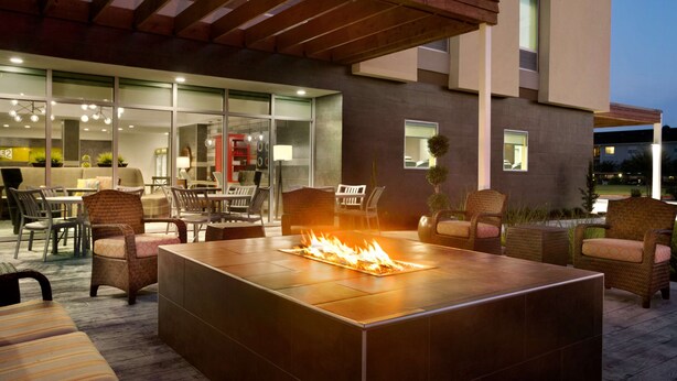 An outdoor patio with a fire feature, seating, and a modern building in the background. - Home2 Suites by Hilton Houston Webster (Webster)