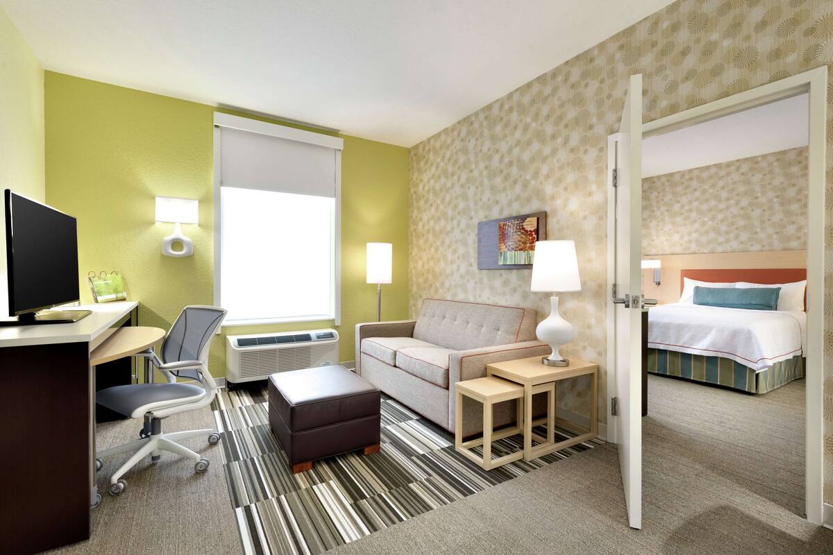A hotel room with a desk, chair, sofa, ottoman, bedside tables, lamp, and a bed.
