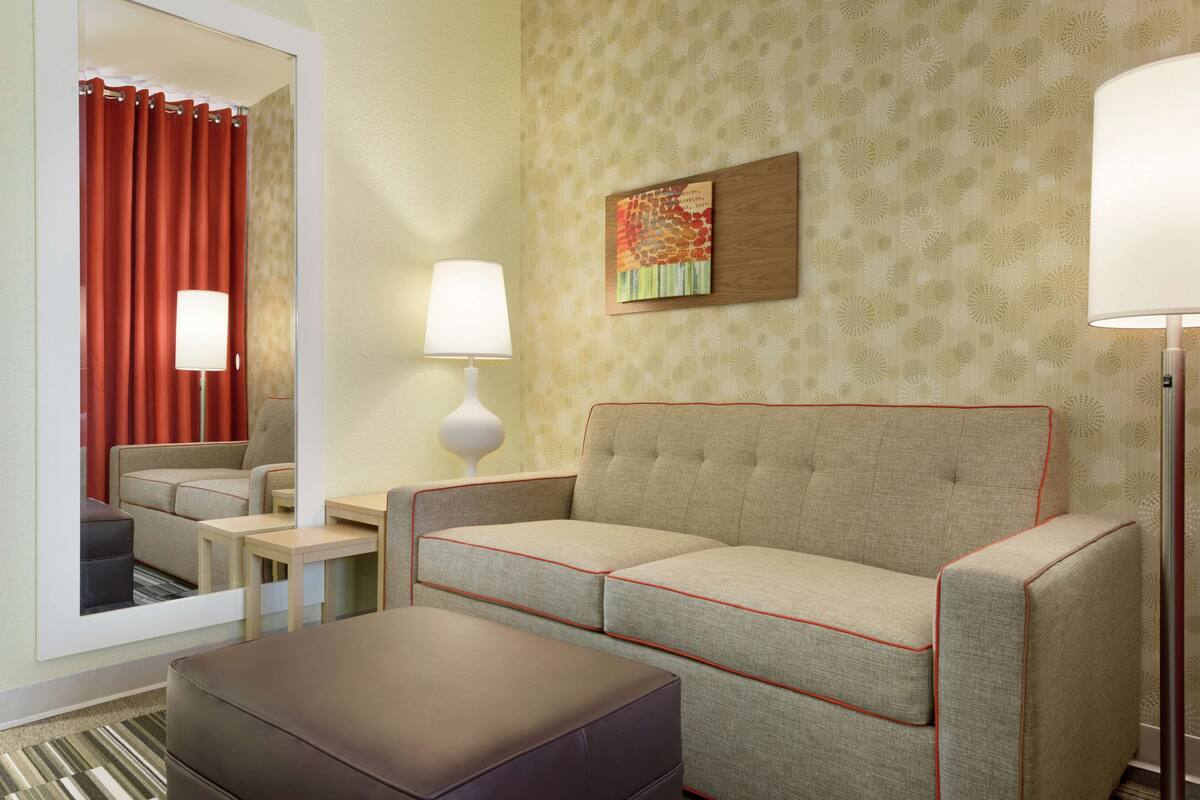 A hotel room with a sofa, ottoman, two lamps, a mirror, and a wall with a patterned wallpaper.