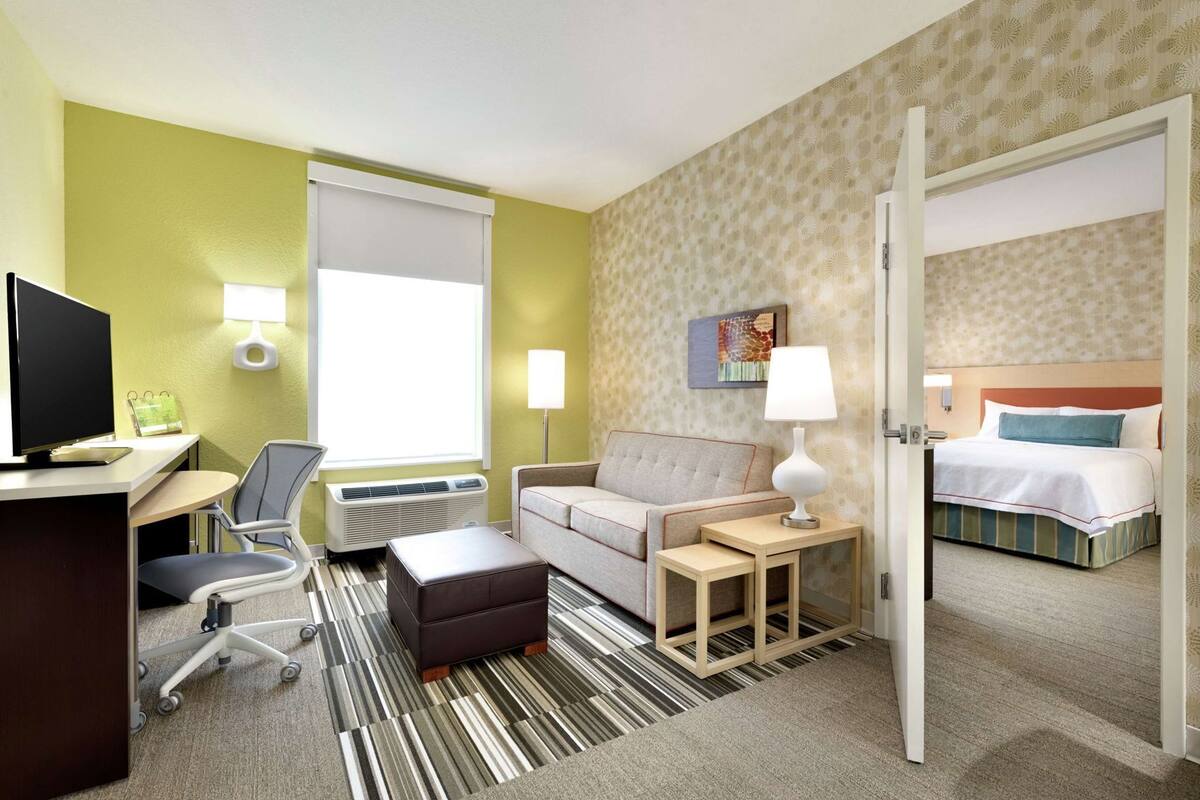 A hotel room with a desk, chair, sofa, bedside table, lamp, and a bed.