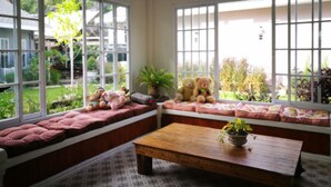 A cozy room with large windows, a wooden bench with cushions, a small table, and a potted plant. - The Goodville Cottage (Pak Chong)