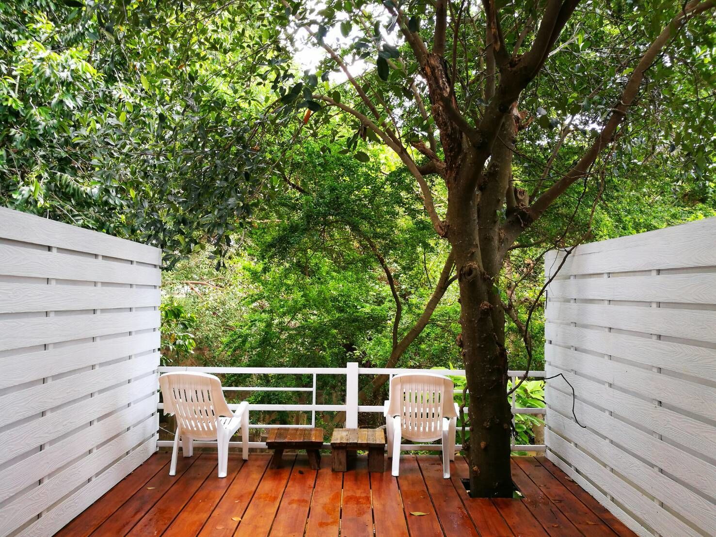 A wooden deck with white plastic chairs and a bench, surrounded by white walls and a tree.