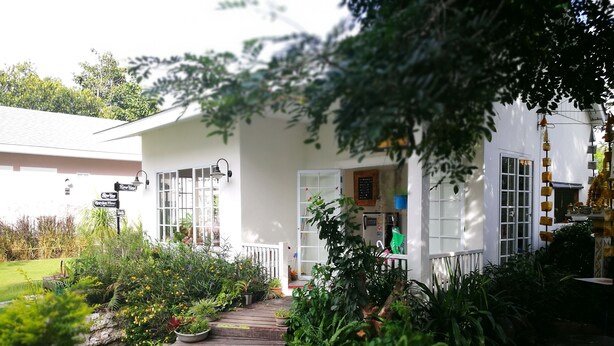 A white building with a covered porch, surrounded by lush greenery and a wooden pathway. - The Goodville Cottage (Pak Chong)
