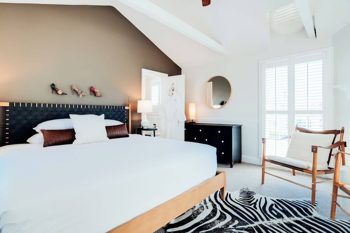 Une chambre à coucher moderne avec un grand lit, une tête de lit en bois, un tapis noir et blanc, une chaise en bois et un miroir au mur.
