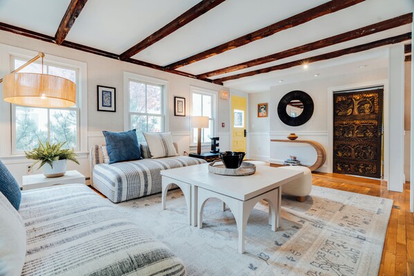 A spacious living room with a high ceiling, exposed wooden beams, a white sofa set, a coffee table, and a large window providing natural light.