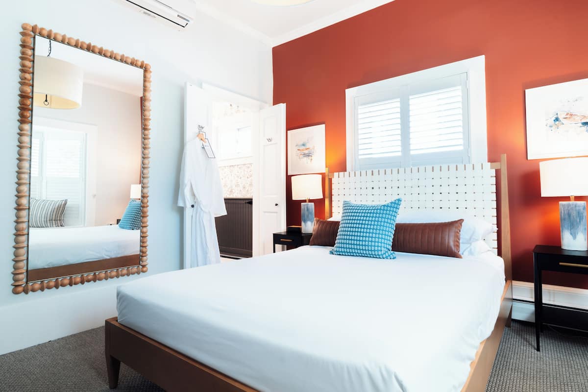 Une chambre d’hôtel avec un grand lit, un miroir à cadre en bois, une fenêtre avec des stores et une table de chevet avec une lampe.