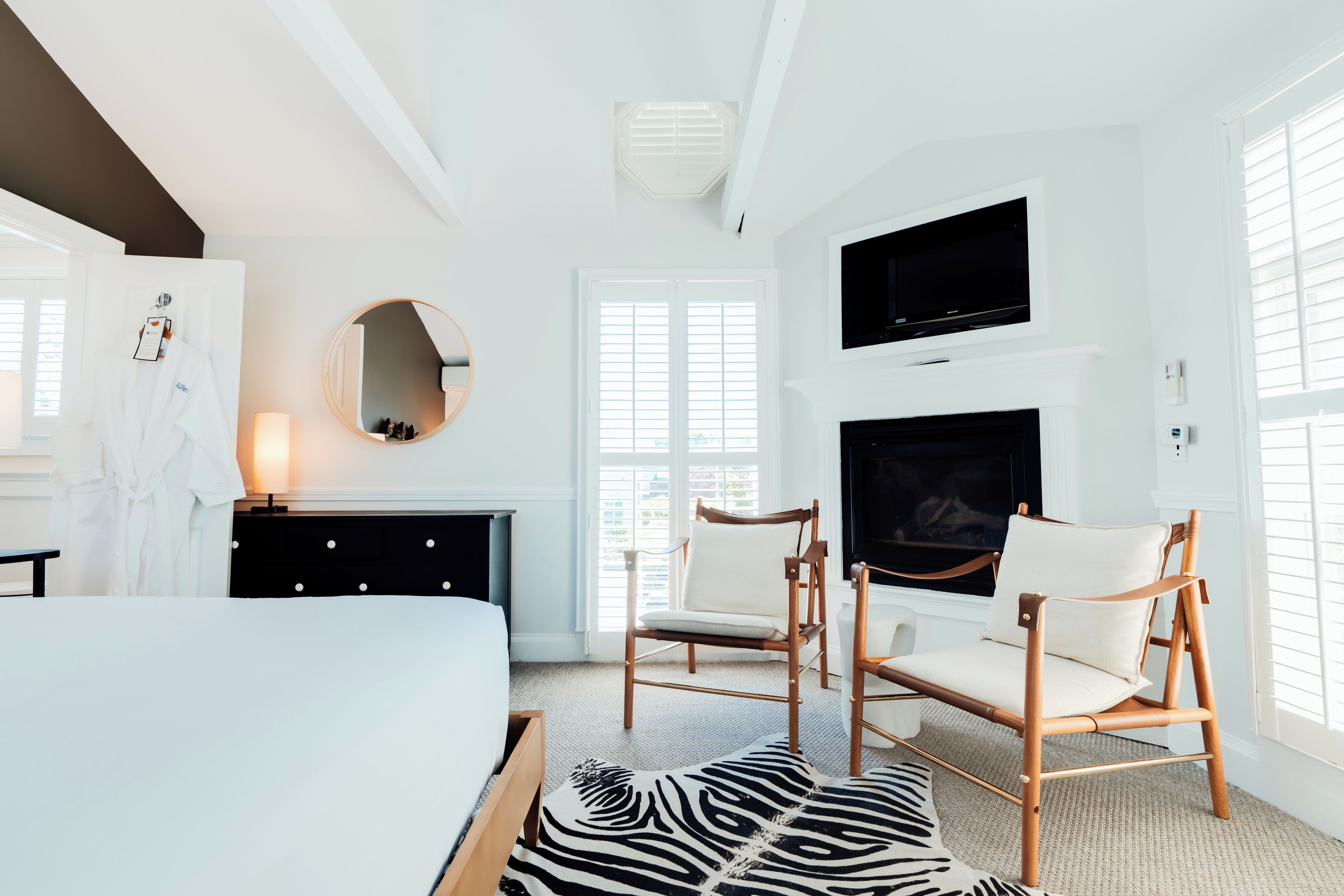 A modern hotel room with a bed, two armchairs, a fireplace, a TV, and a large window with blinds.