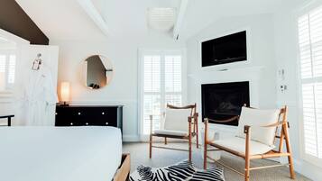 A modern hotel room with a bed, two armchairs, a fireplace, a TV, and a large window with blinds.