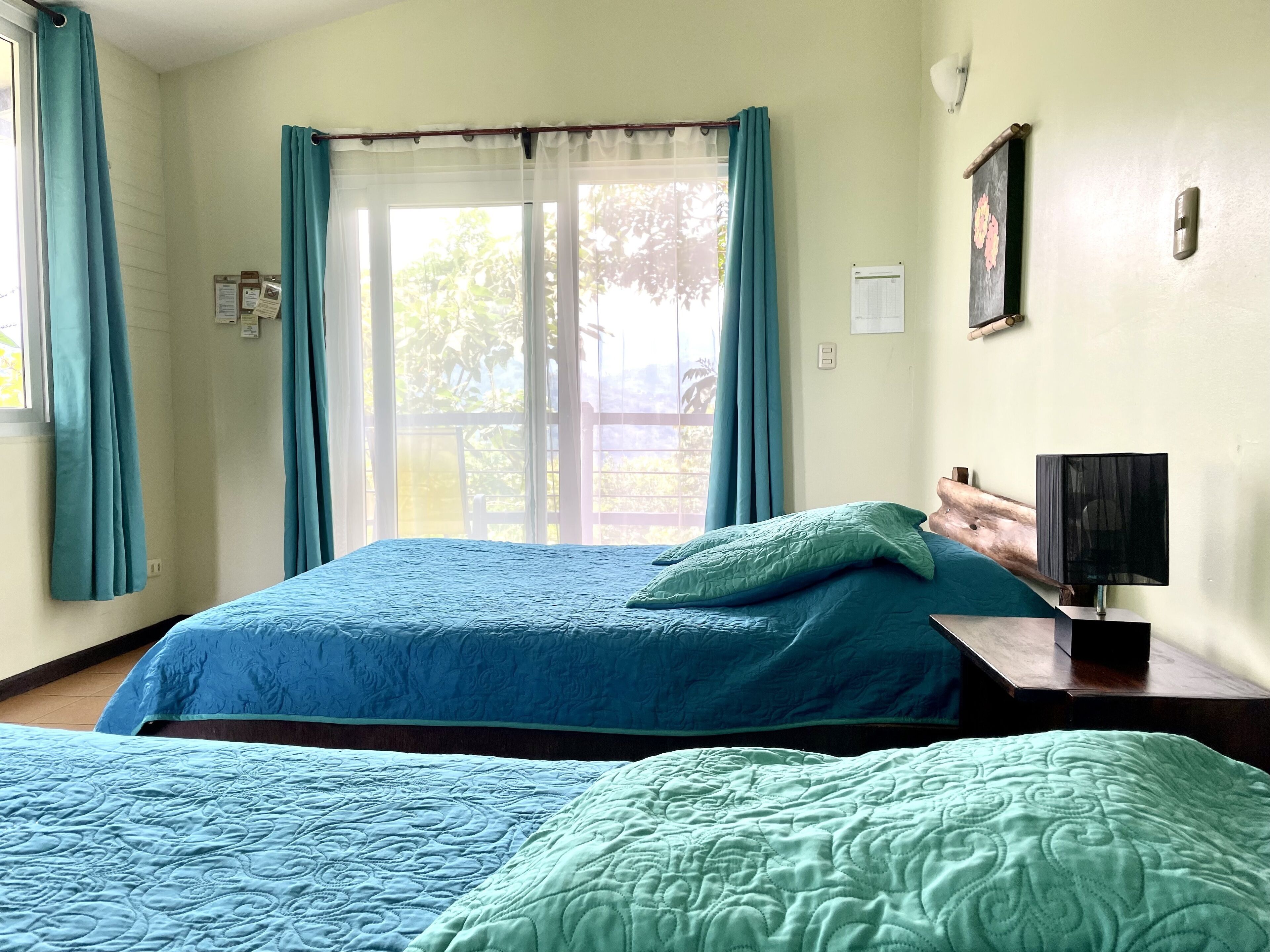 Una habitación con dos camas, una ventana con cortinas, una mesita de noche con lámpara y una luminaria en la pared.