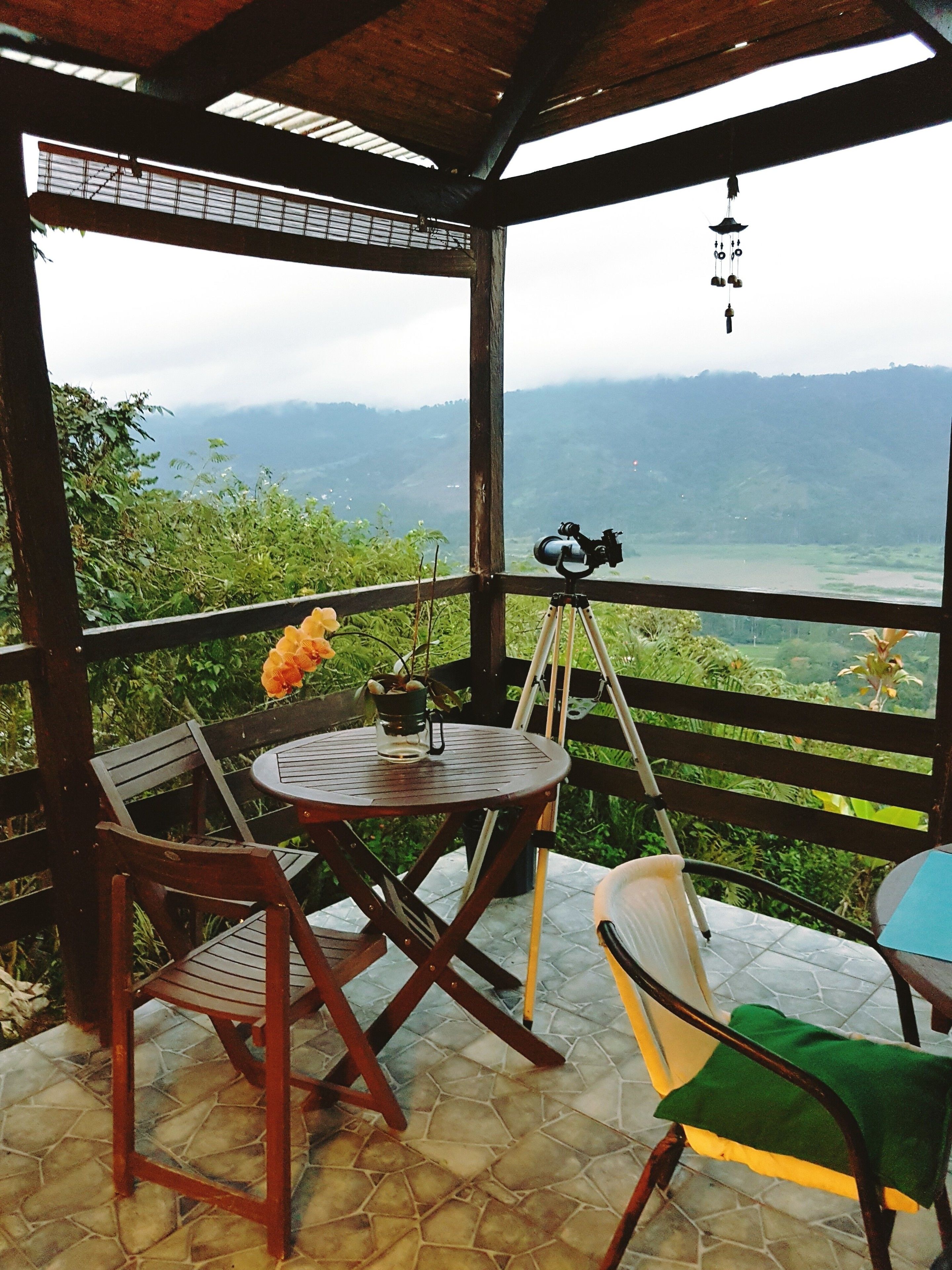 A covered outdoor seating area with a table and chairs, a telescope, and a view of a mountainous landscape.