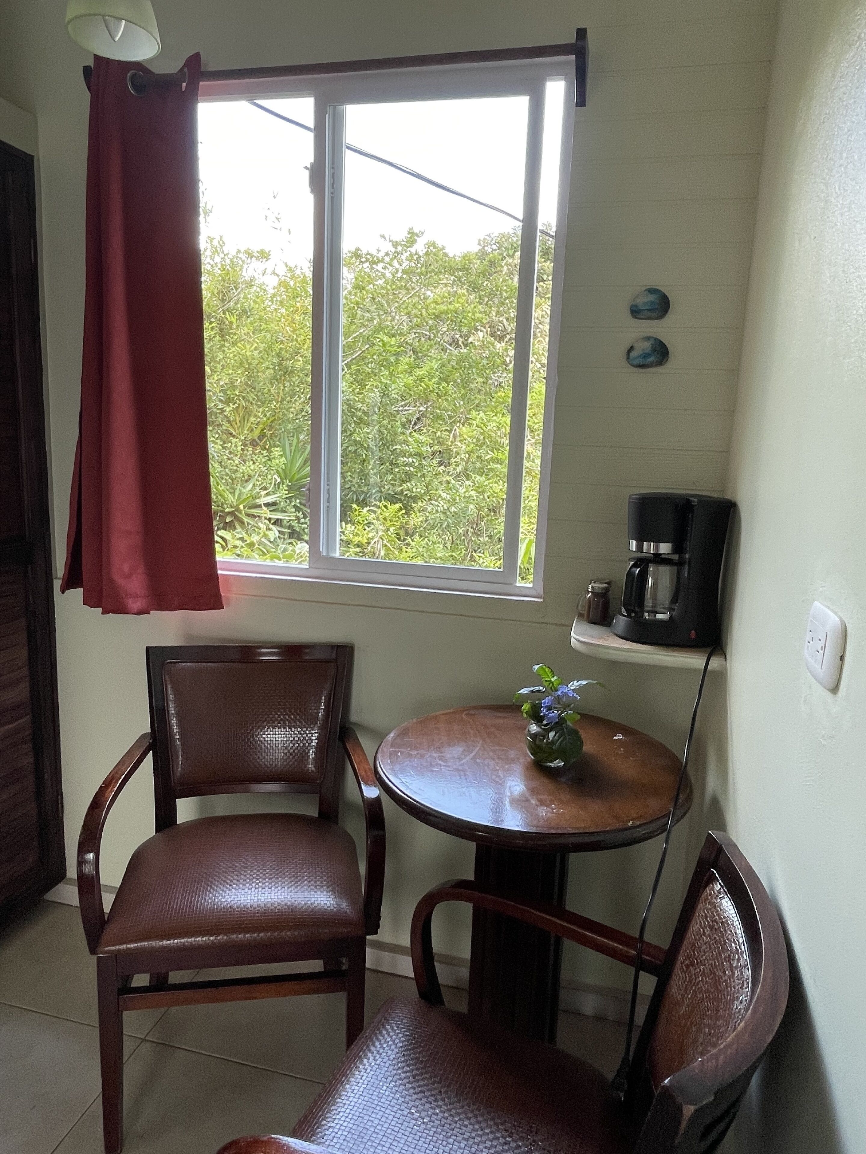 A small room with a window, two chairs, a small table with a vase, and a coffee maker on a shelf.