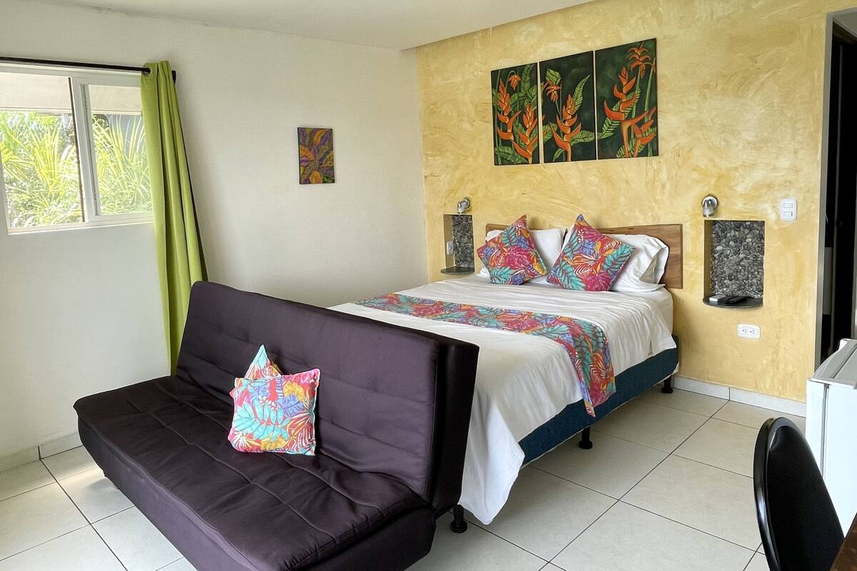 A bedroom with a bed, a sofa, a small table, and a wall-mounted shelf.