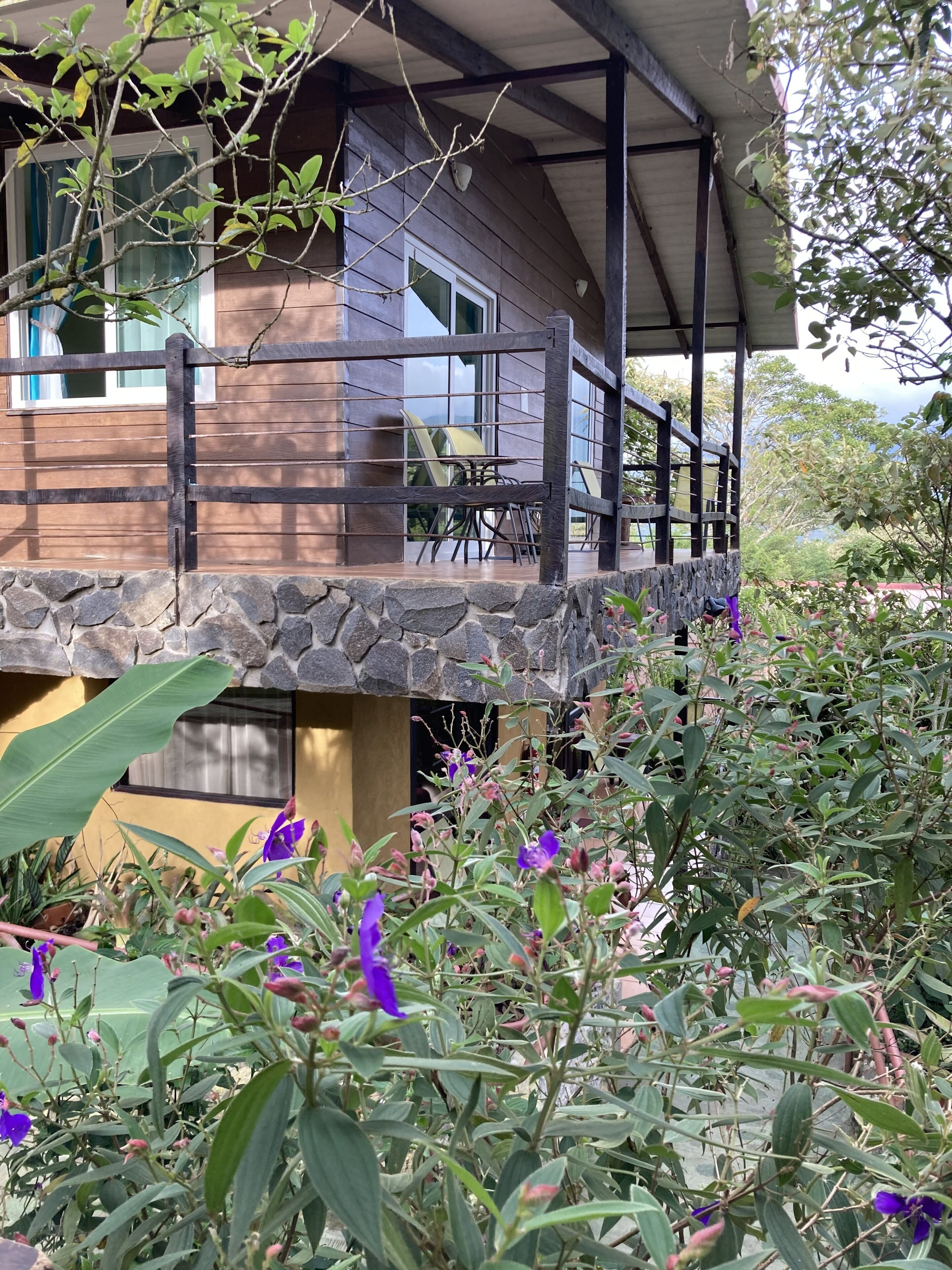 A wooden house with a stone foundation, a balcony with chairs, and a garden with purple flowers.