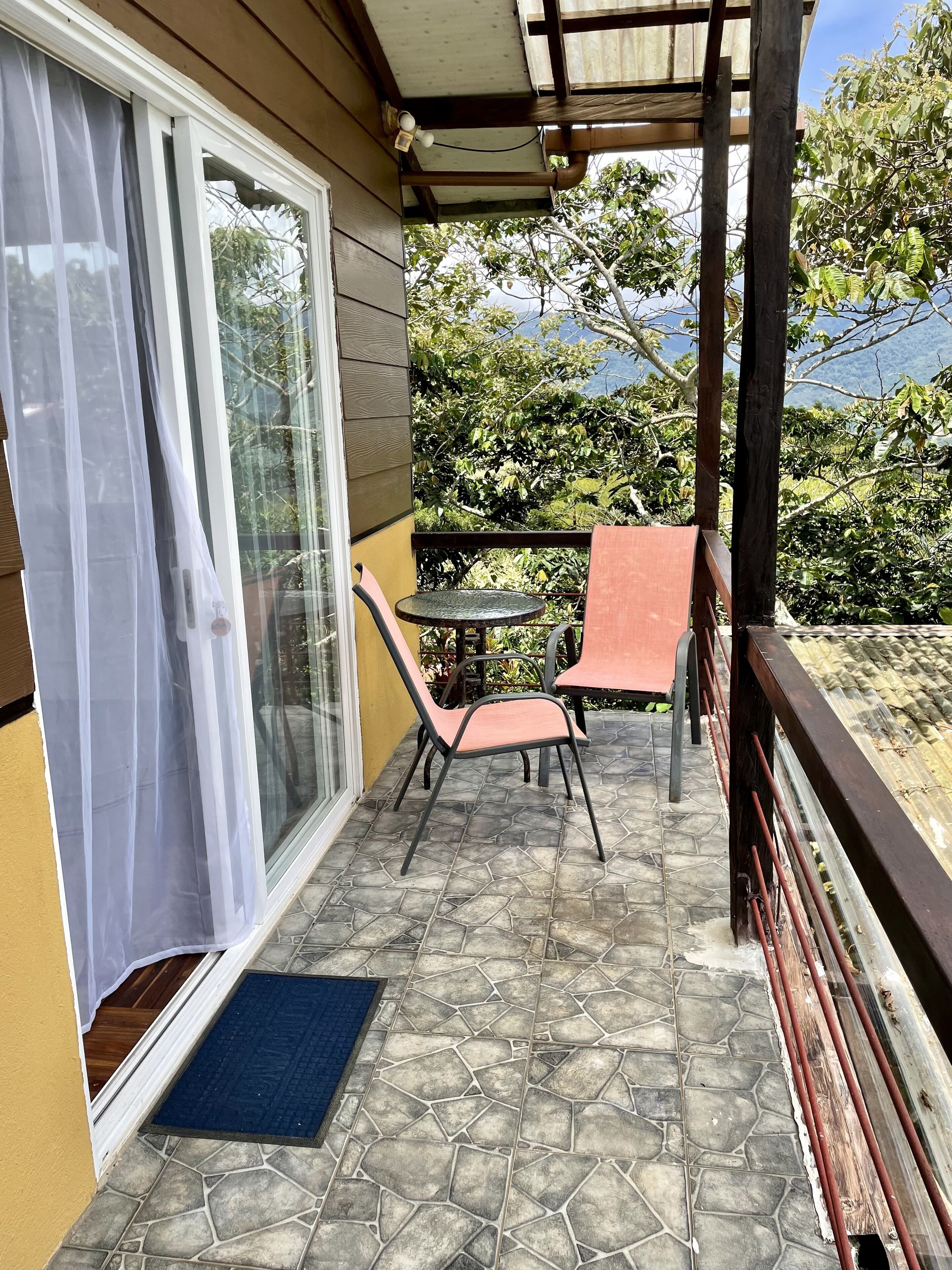 A balcony with a table and chairs, a blue mat, and a view of greenery and mountains.