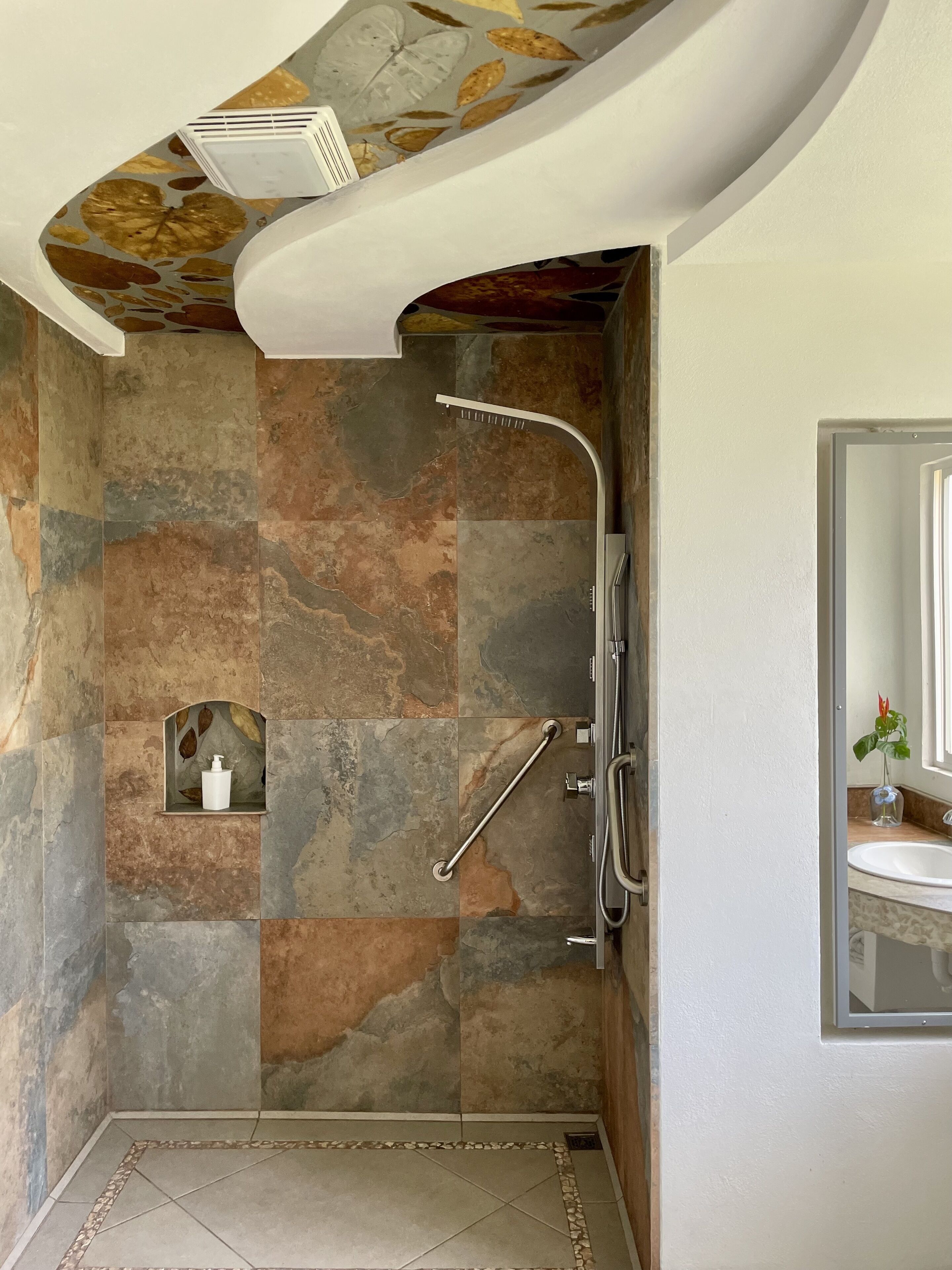 Un baño con pared de piedra, ducha, barra de apoyo y lavabo con espejo.