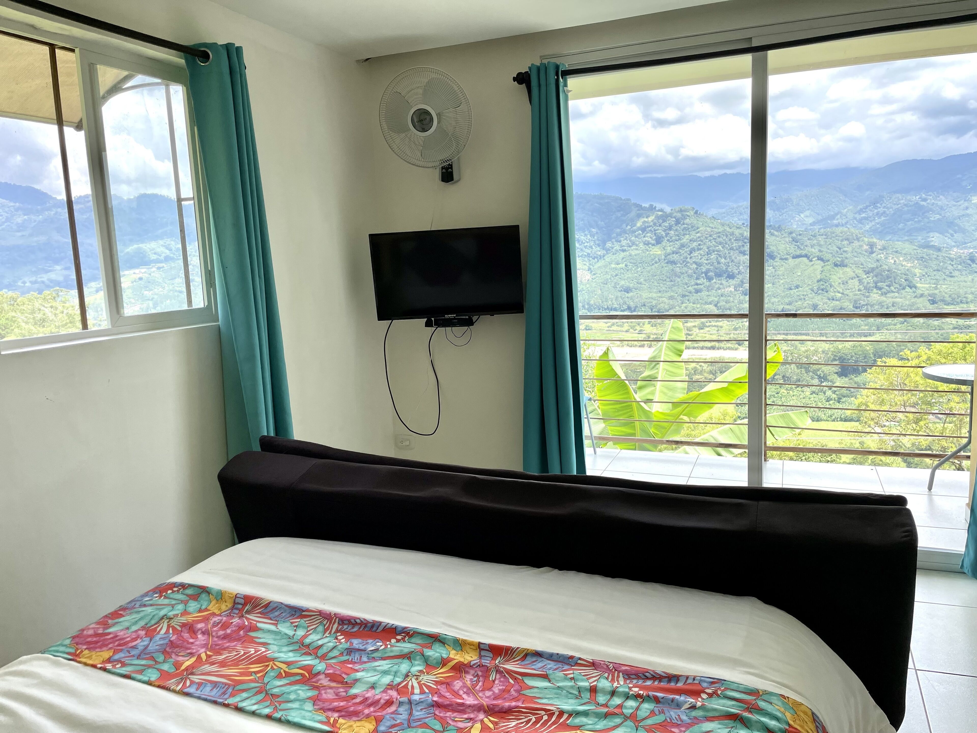Habitació d'hotel amb un llit, una televisió i un balcó que ofereix vistes a muntanyes i vegetació.