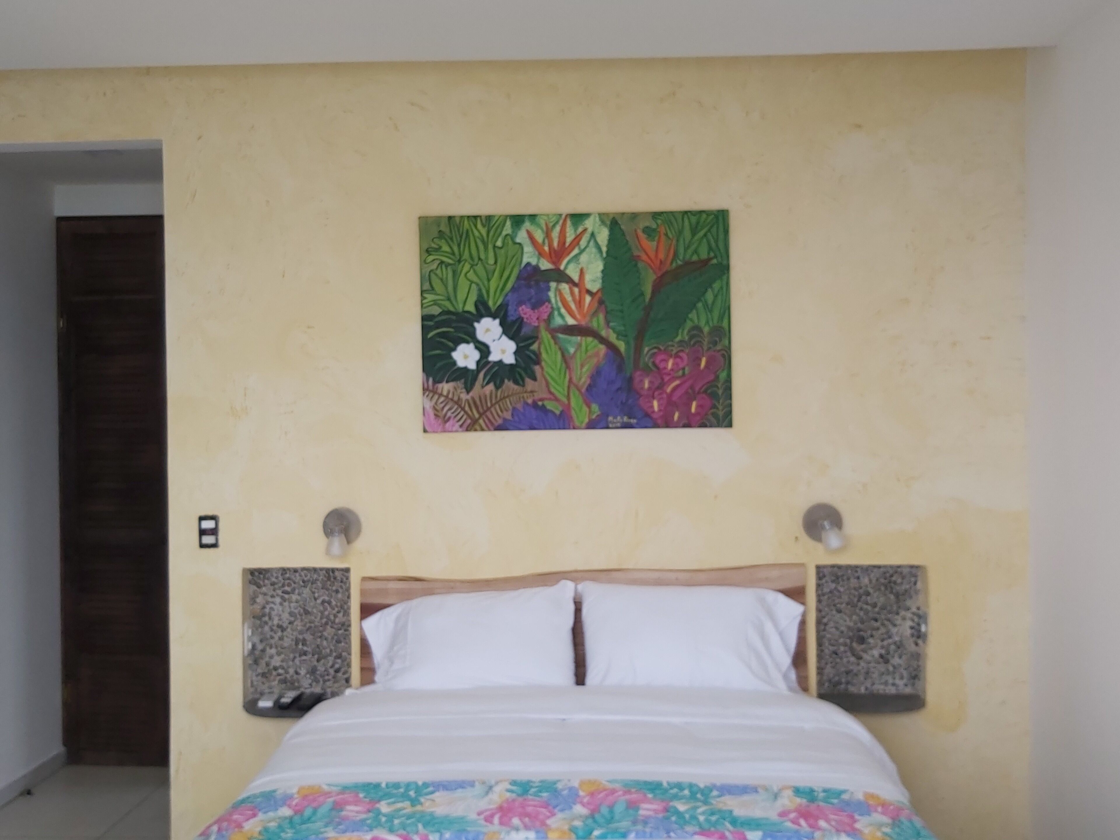 A bedroom with a bed, white pillows, a floral bedspread, a wall-mounted painting, and a door.